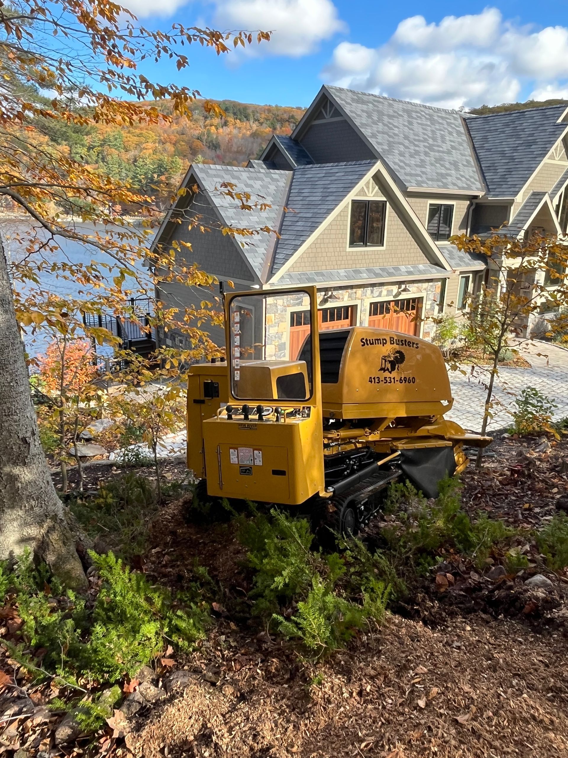 Yellow stump grinder in front of a stone house with a lake view, fall foliage.