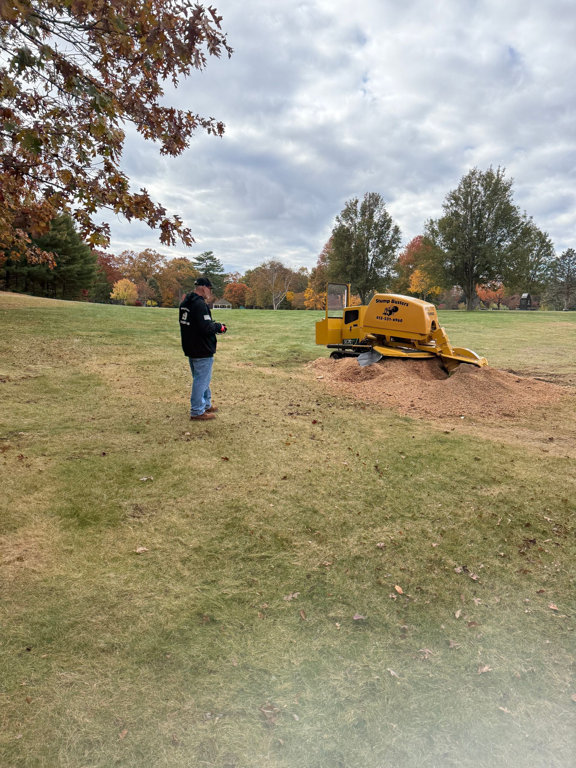A person operates a yellow stump grinder on a grassy field on a cloudy day.