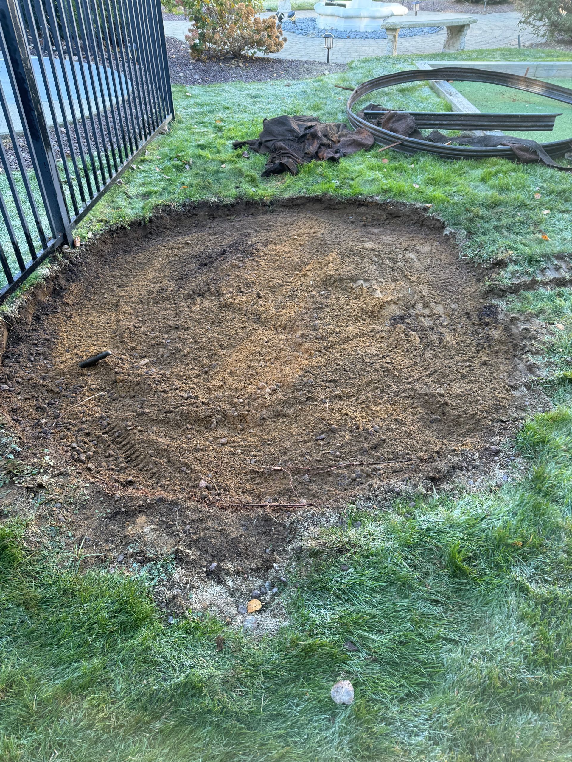 A circular area of dirt in a grassy yard, near a black fence, with some construction.