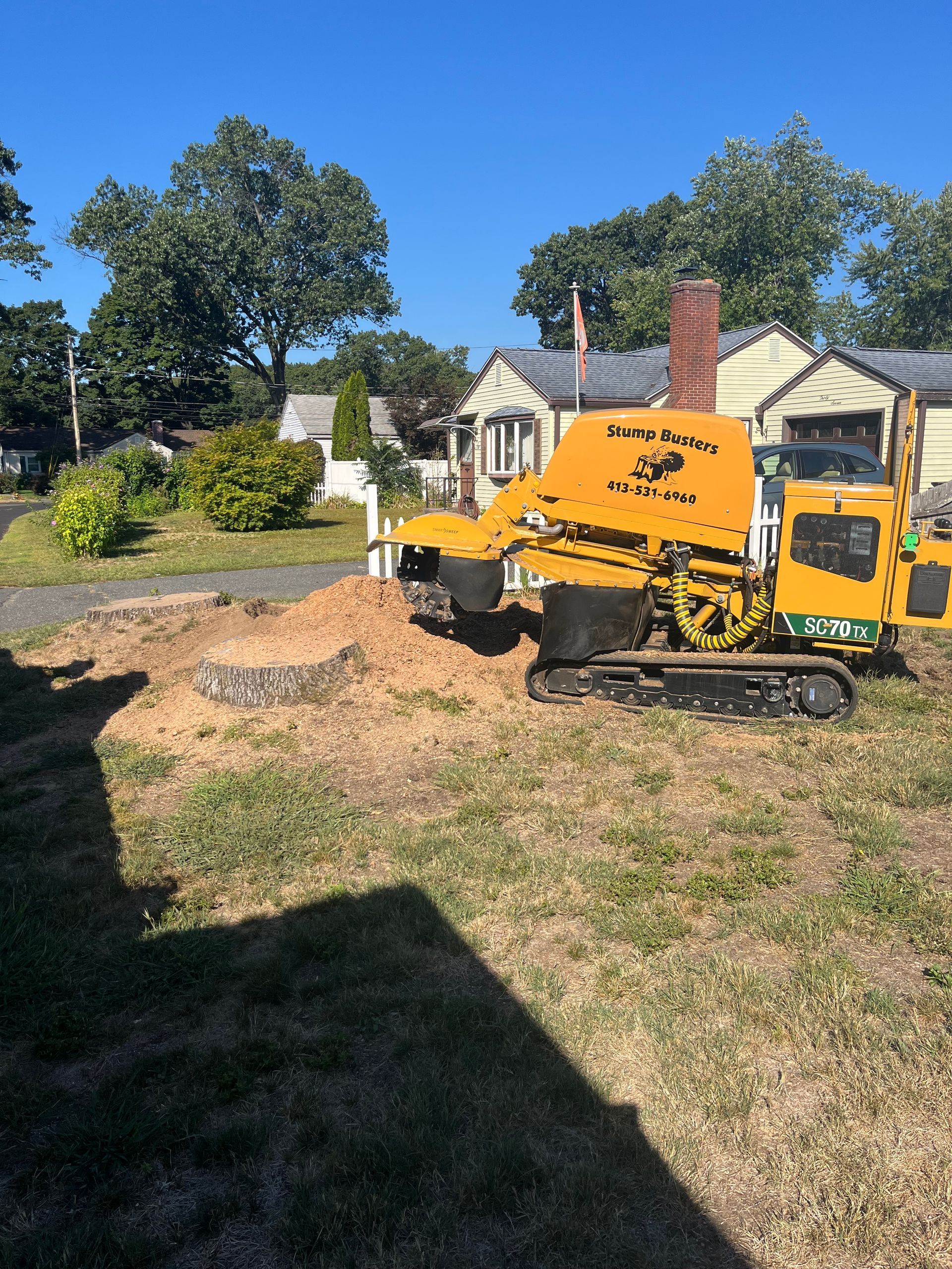 Yellow stump grinder on tracks, grinding a stump in a residential yard, sunny day.
