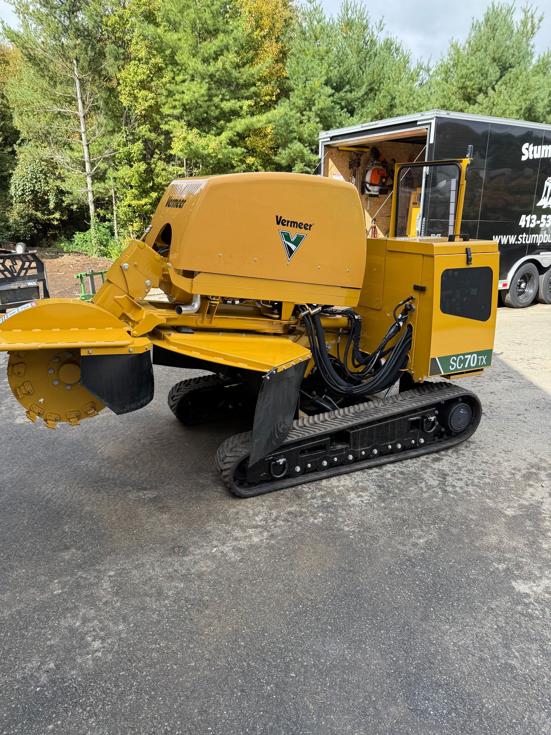 Yellow and black stump grinder on tracks, parked on pavement, next to a trailer.