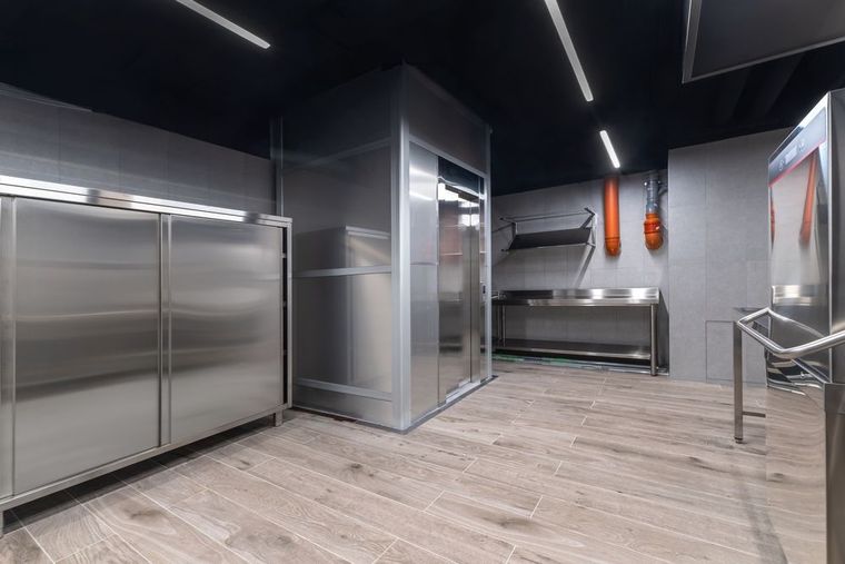 Modern stainless-steel commercial kitchen with bright overhead lights and light wood-look flooring