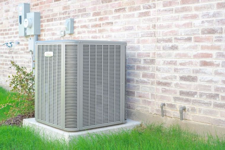 Outdoor air conditioning unit beside a brick wall on a concrete pad, with grass and shrubs nearby