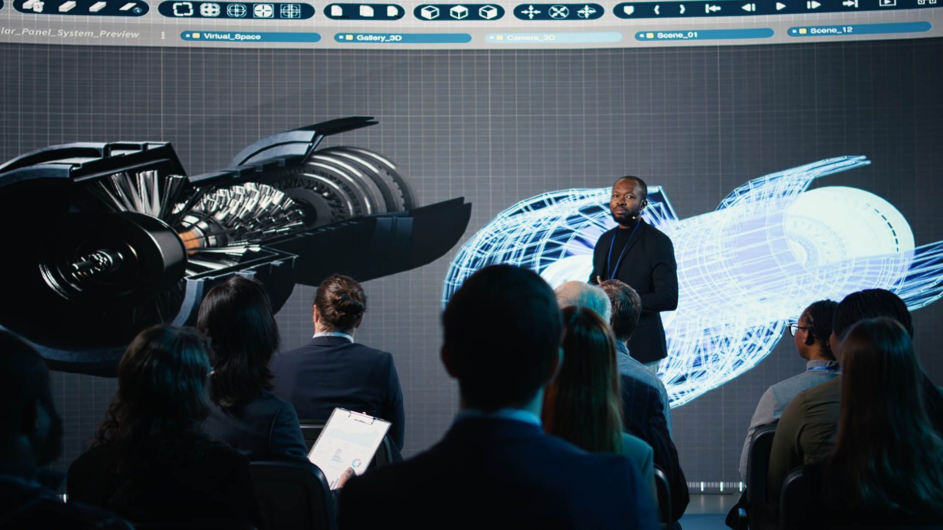 Man presenting design schematics to an audience. A large screen displays a 3D model.