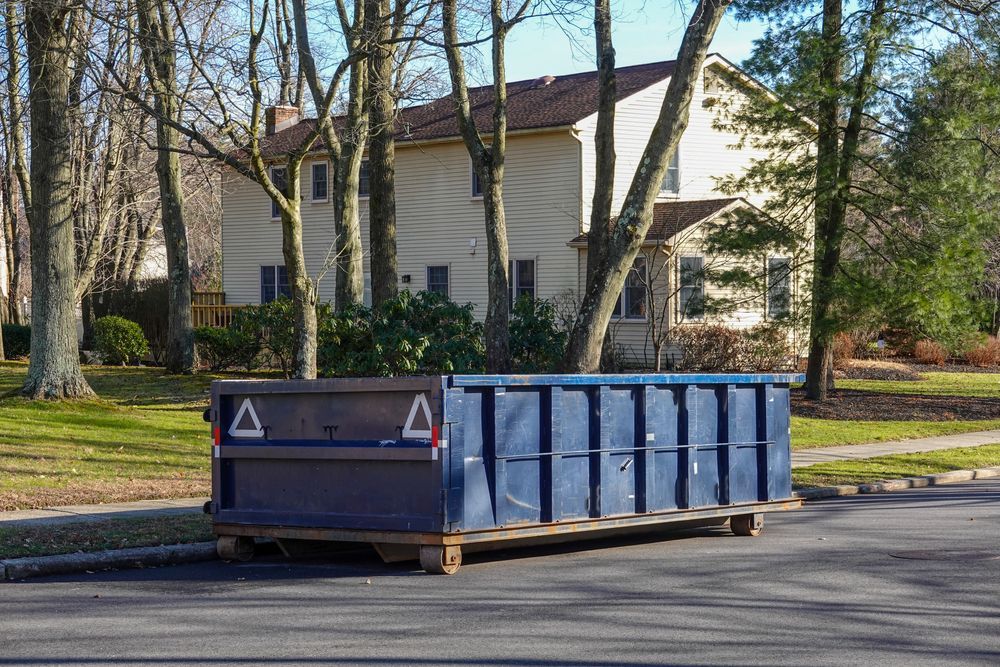 Blue dumpster on a residential street; house in the background, trees along the curb.