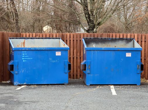 Two blue dumpsters next to each other in a parking lot, in front of a wooden fence.