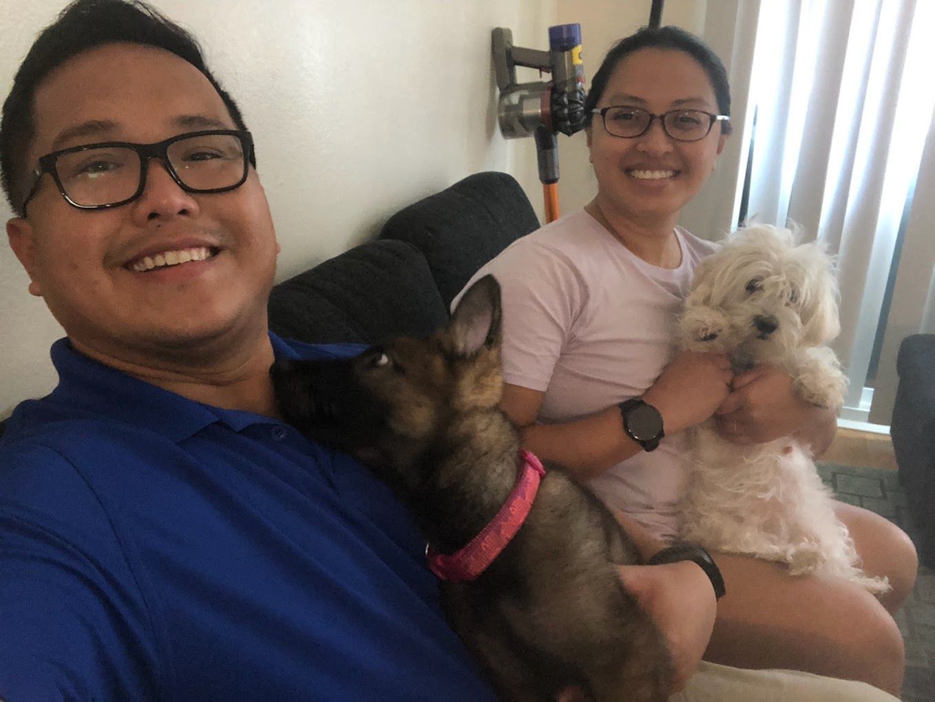 A couple smiles while holding two dogs on a couch; the man wears glasses and blue shirt.