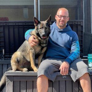 Man with shaved head and dog posing outdoors, both smiling, sitting on wooden box.