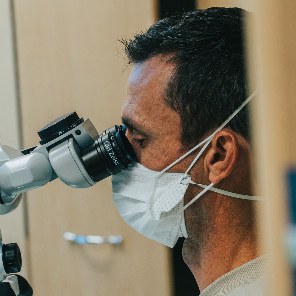 Person wearing a mask looks through a dental microscope with a Zeiss lens in a medical office.