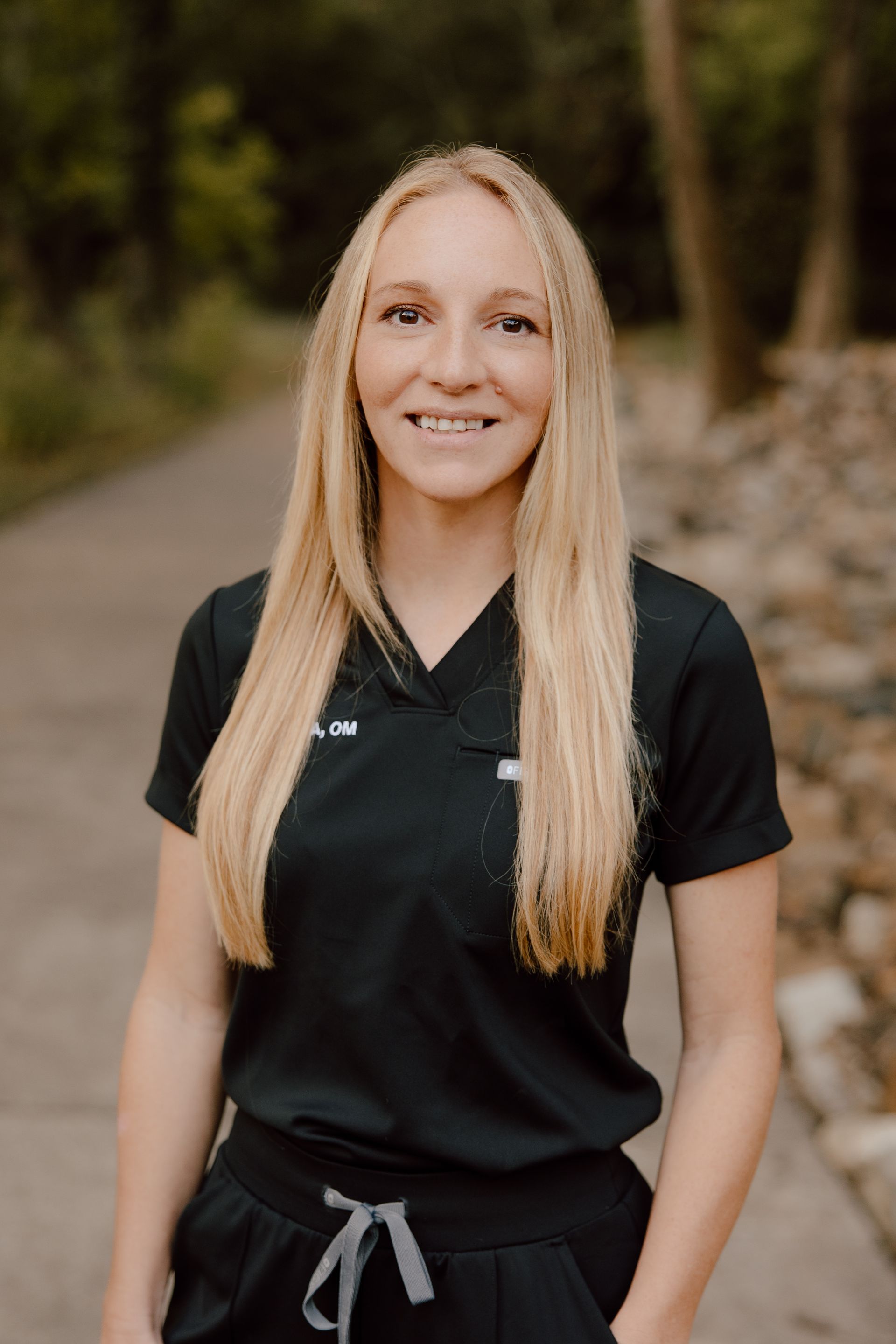 Blond woman in black scrubs smiles at the camera on a path with trees.