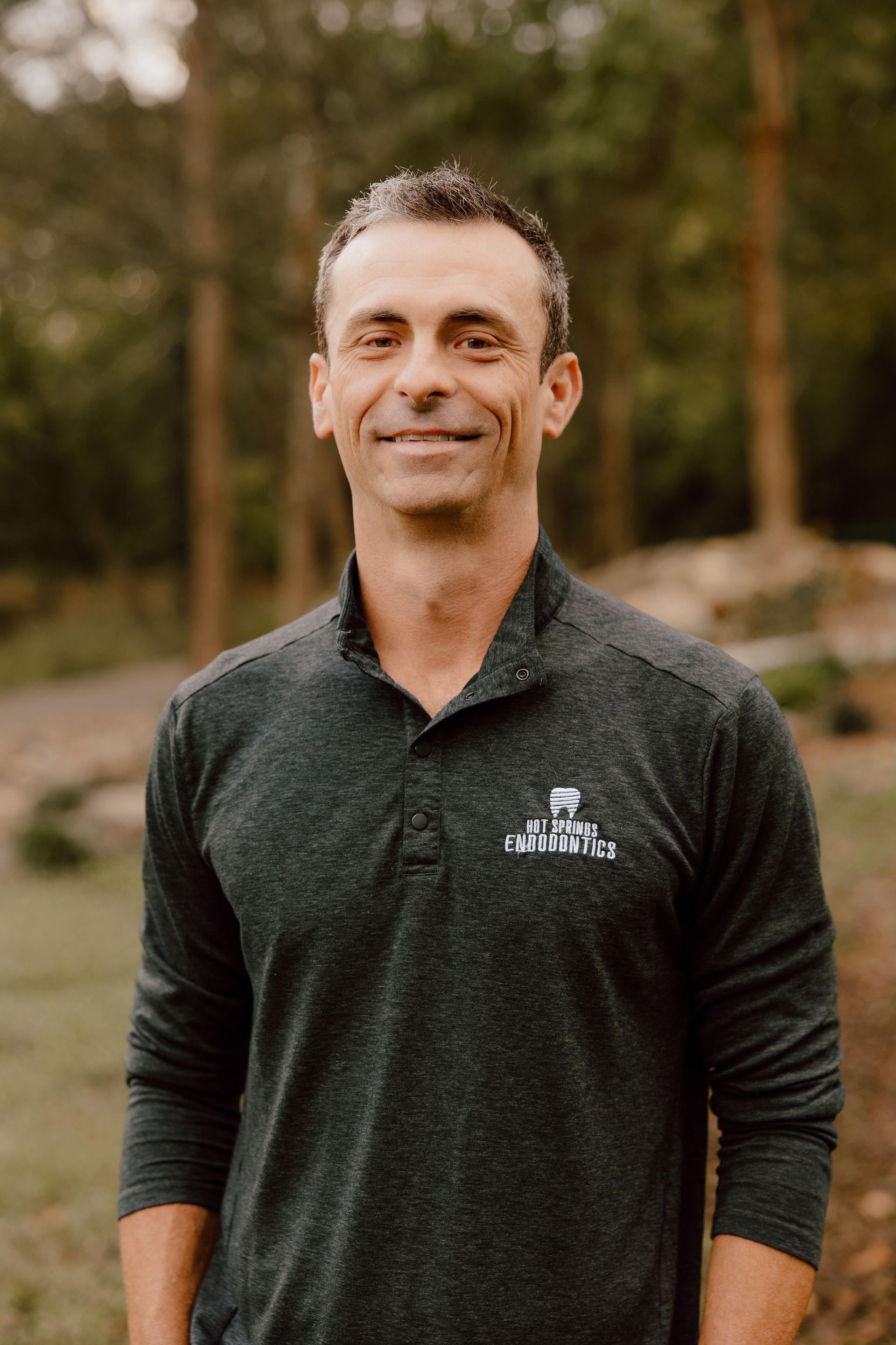 Man in gray shirt smiles, standing outdoors in front of trees.