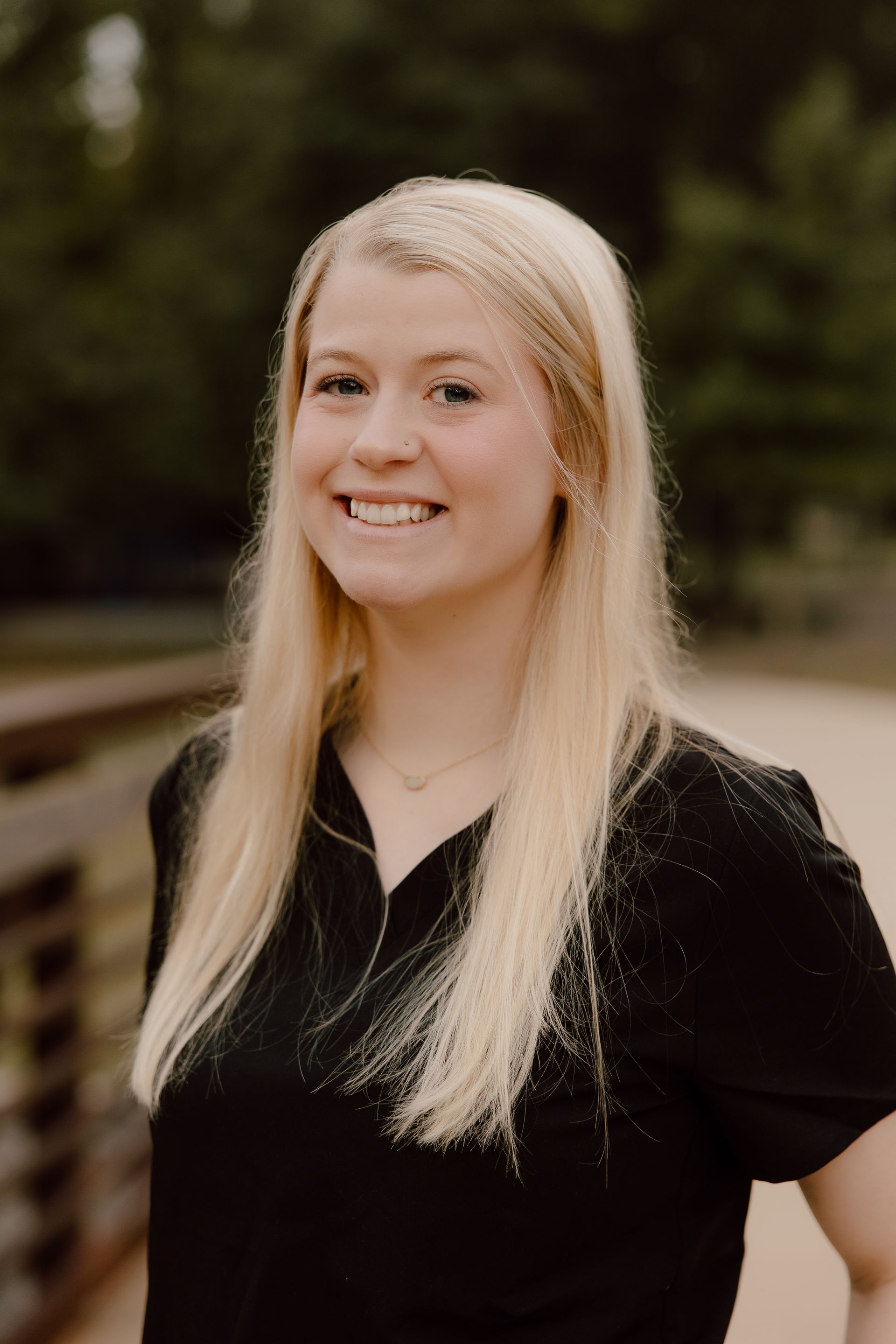 Blond person smiling, black shirt, outdoor setting, bridge visible.