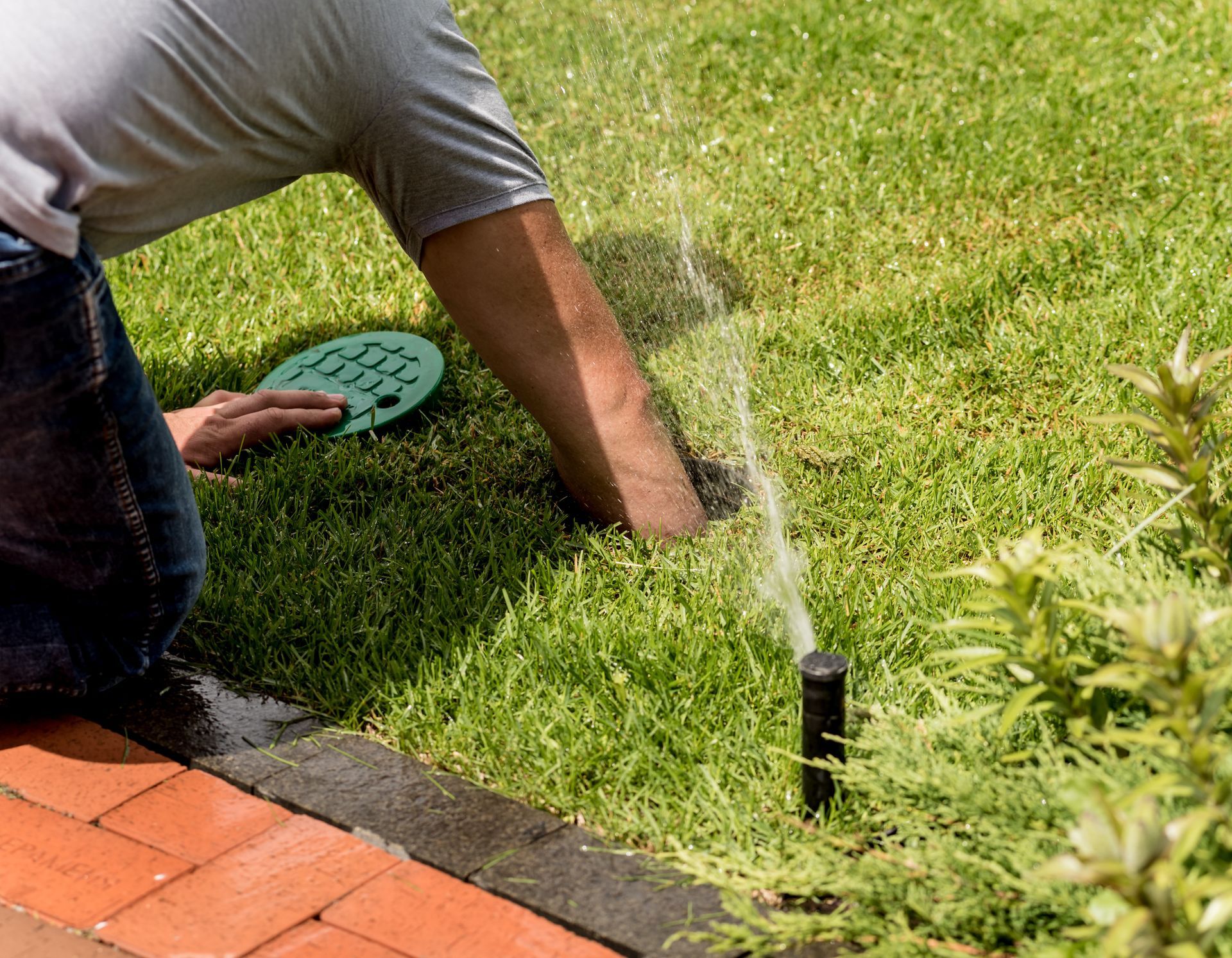 Person kneeling on lawn repairs a black sprinkler head near a brick patio