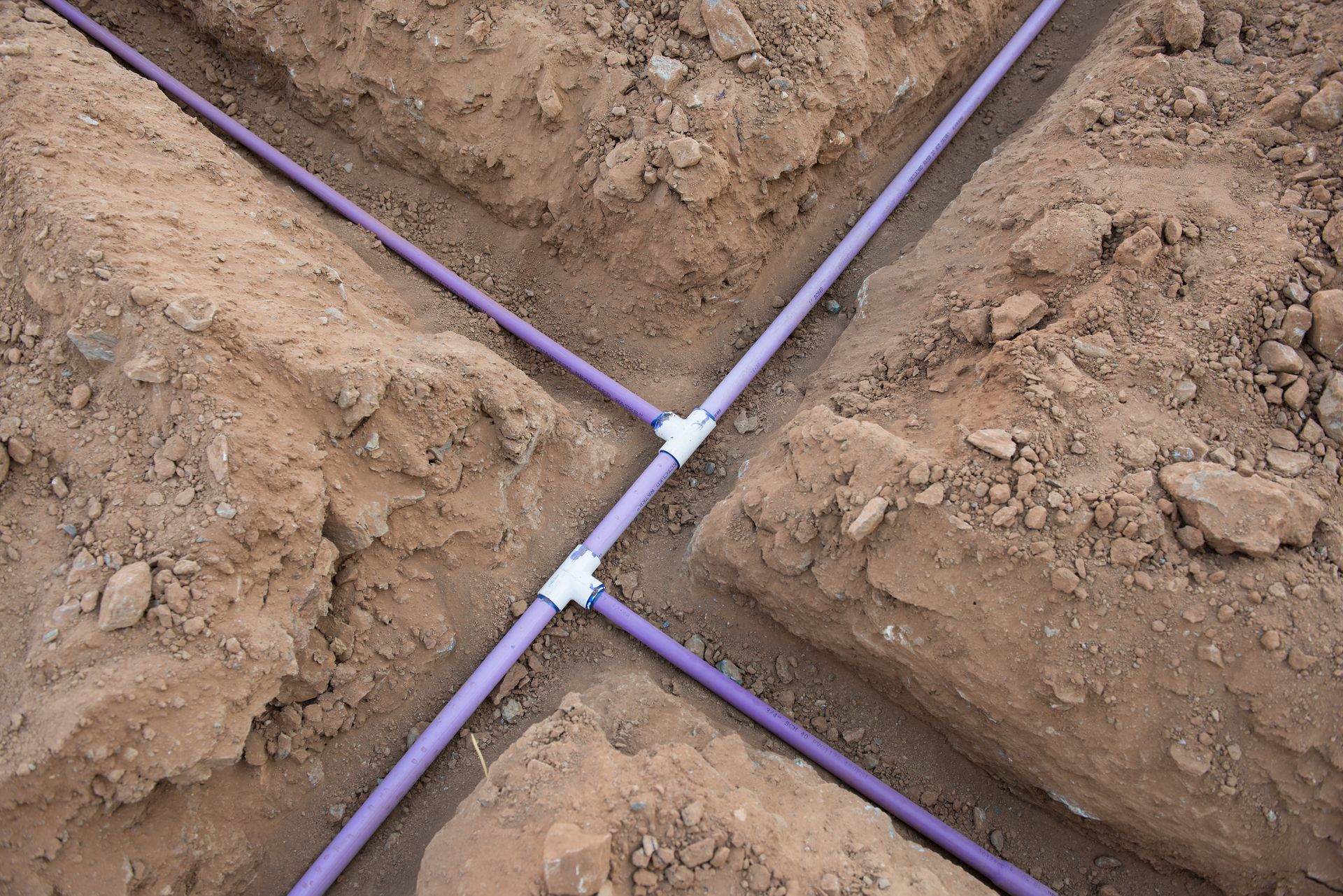 Purple irrigation pipes branching through a dirt trench, forming a cross junction in soil rows