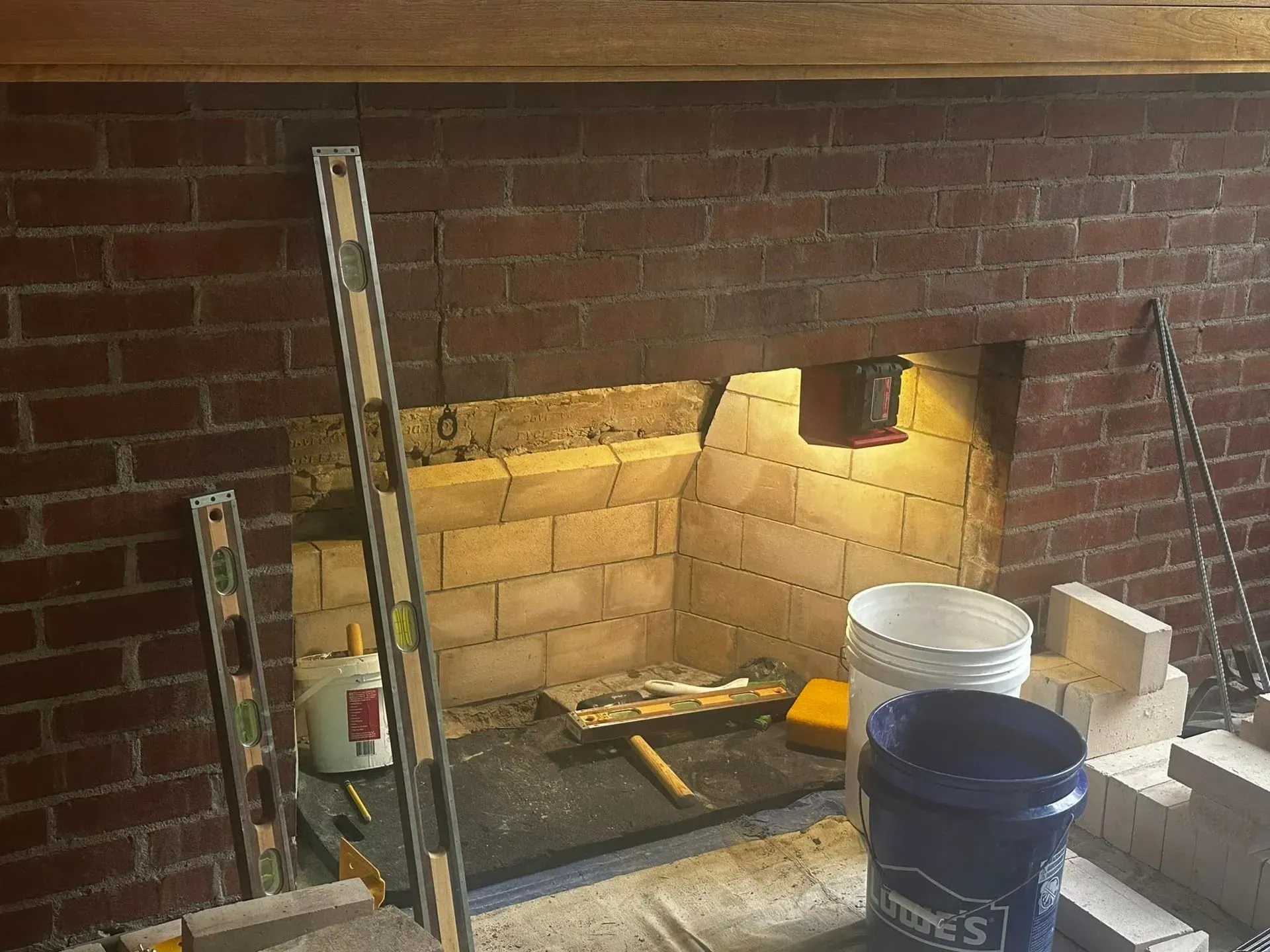 Fireplace under construction, showing exposed brick and firebricks, with tools and buckets.