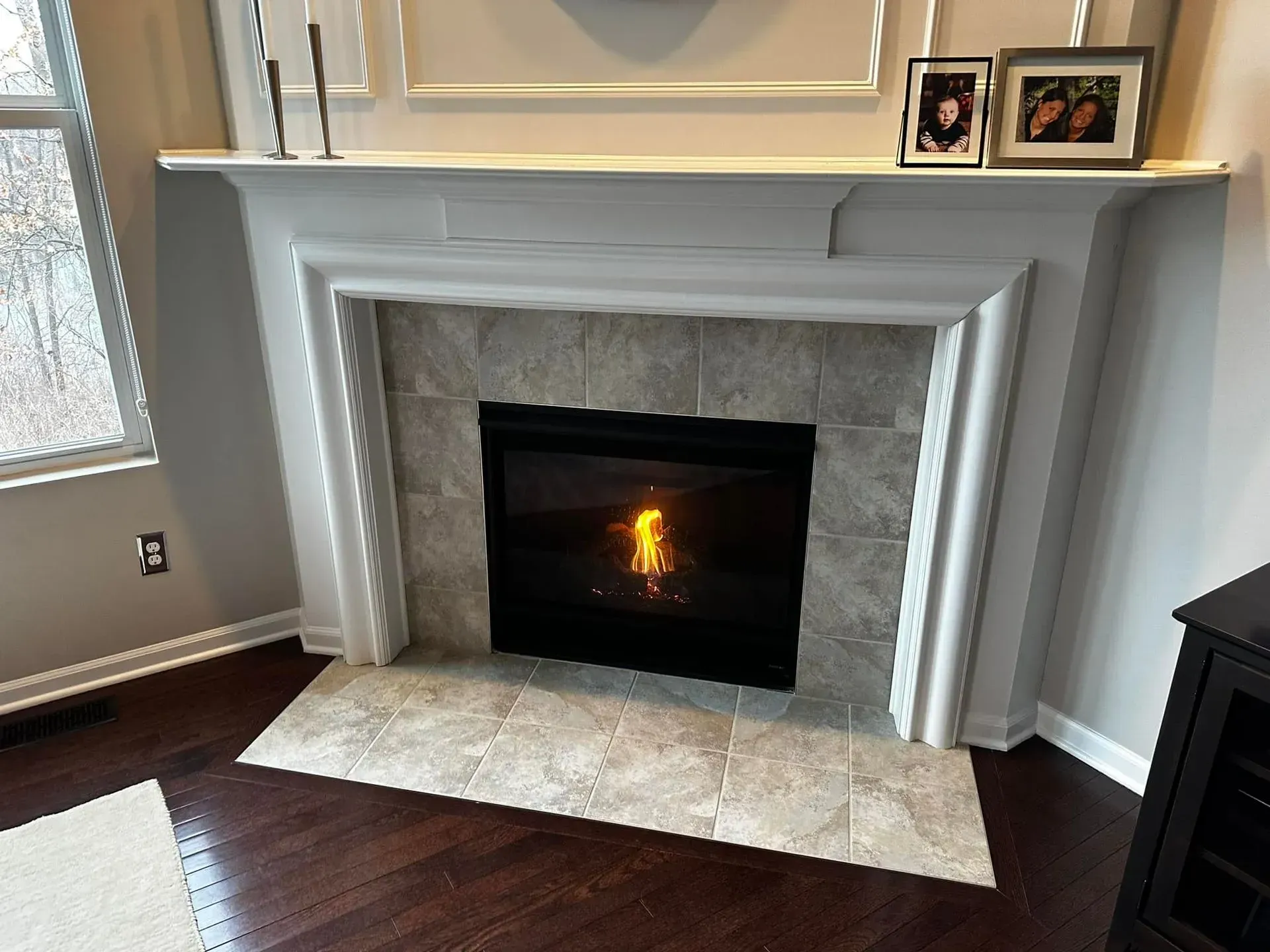 Fireplace with lit flames, surrounded by gray tile and white mantel. Dark wood floor.