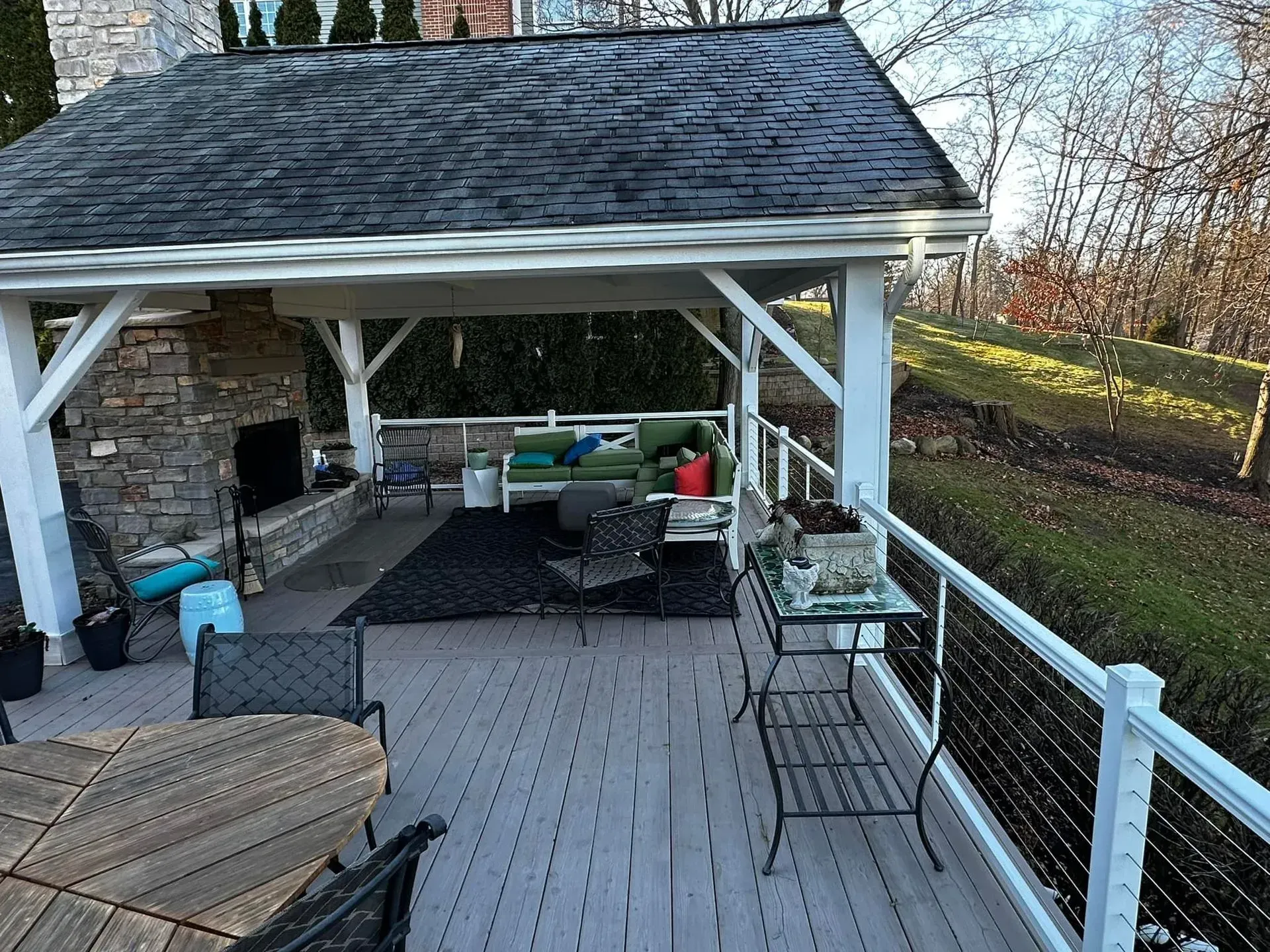 Outdoor covered patio with stone fireplace, seating, and a railing overlooking a yard.