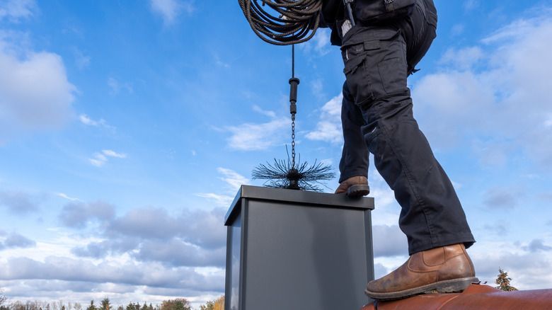 A person on a metal roof sweeps a brick chimney with a long wire brush against a cloudy sky.