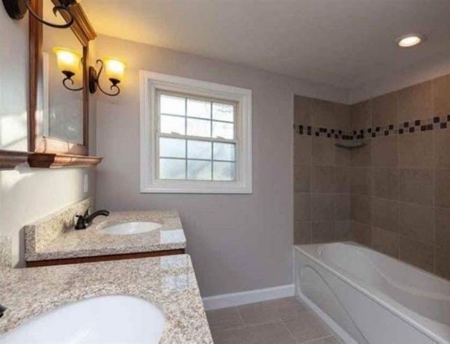 Bathroom with tan tiled shower, double sink granite countertop, and small window.