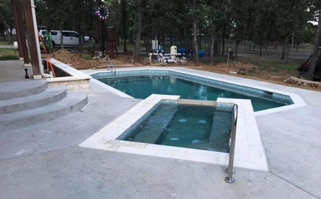 Outdoor pool and attached spa, both with tiled edges, set in concrete, surrounded by trees.