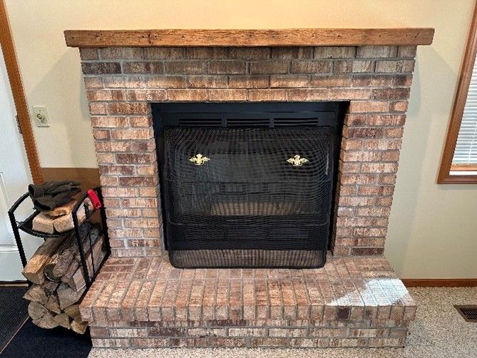 Brick fireplace with black mesh screen and wooden mantel, firewood in a rack.