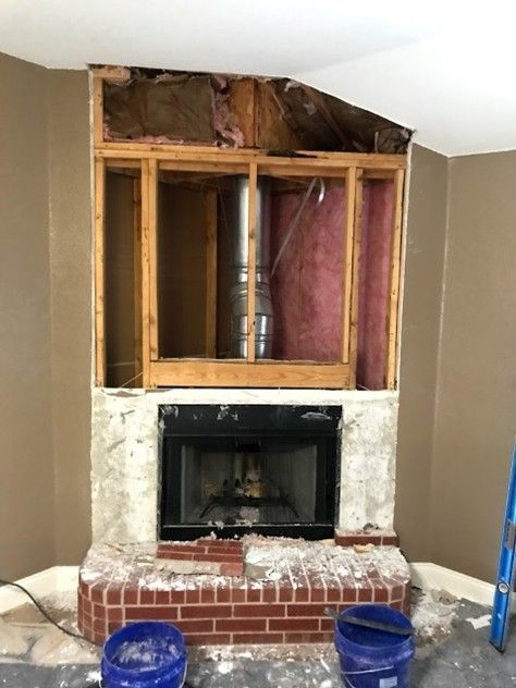 Fireplace under renovation; exposed wooden framing, brick base, and partially demoed walls; two blue buckets.