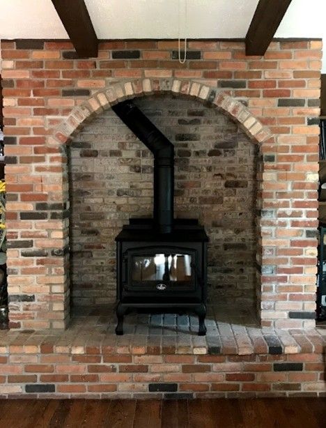Brick fireplace with arched opening housing a black wood-burning stove and chimney. Brown beams overhead.