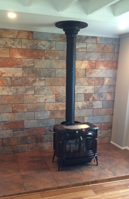 Black wood-burning stove with vertical pipe against a brick-like wall and tiled floor.
