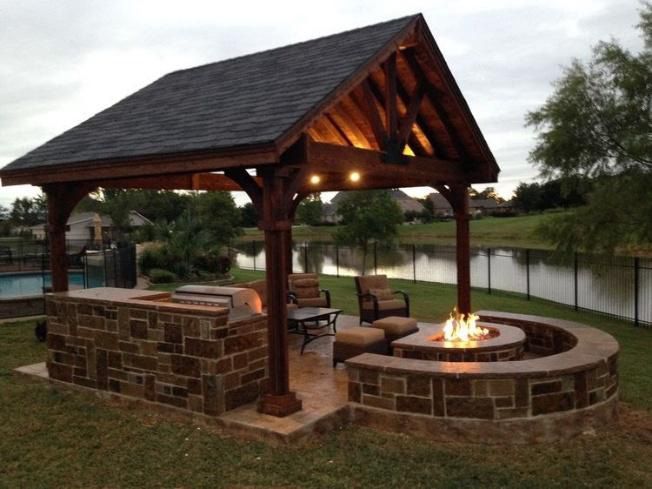 Outdoor structure with a stone fire pit, seating, and a covered cooking area next to a pond.