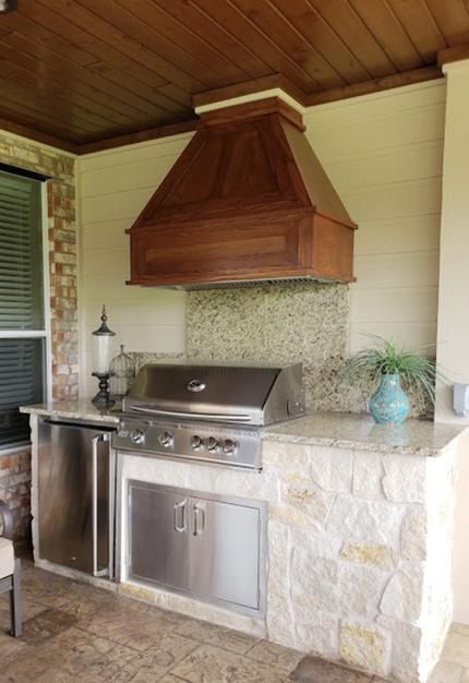 Outdoor kitchen with stainless steel grill, stone countertop, wood hood, and refrigerator.