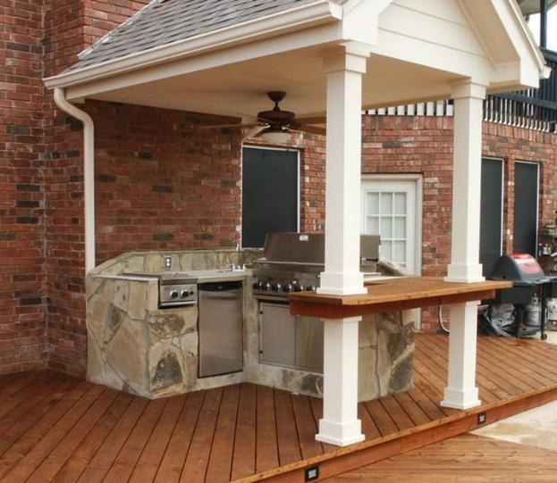 Outdoor kitchen with brick, stone, and wooden surfaces. Includes grill, sink, and bar area, with a fan and deck.