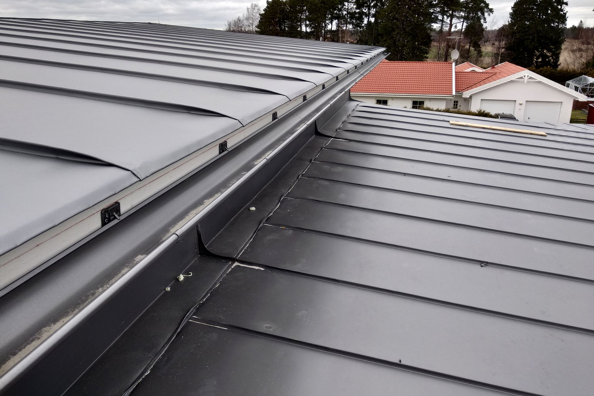 A high-angle view of a gray metal standing seam roof with a central valley flashing separating two sloped roof sections.