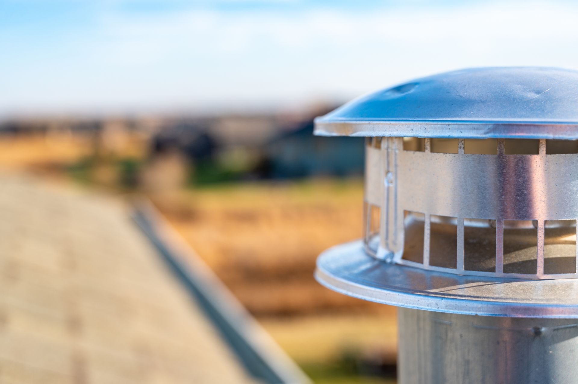 A close-up of a metal chimney cap on a shingled roof, with a blurred residential neighborhood in the background.