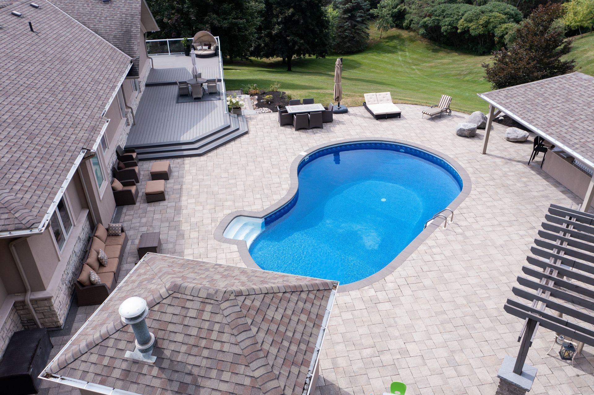 A high-angle view of a backyard with a kidney-shaped pool, stone patio, wooden deck, gazebo, and outdoor furniture.
