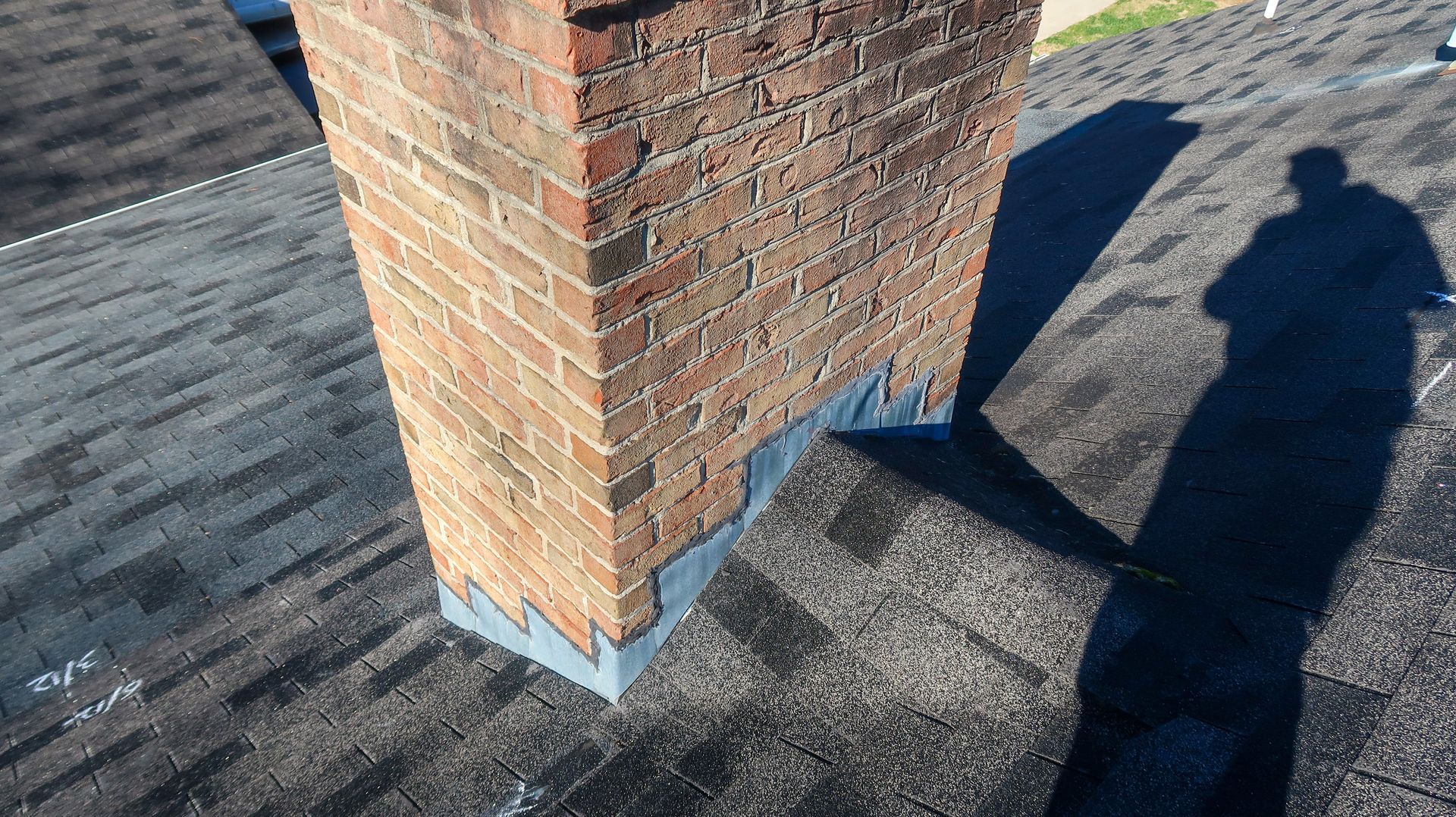 A brick chimney on a shingled roof features metal flashing around its base, with the shadow of a person nearby.