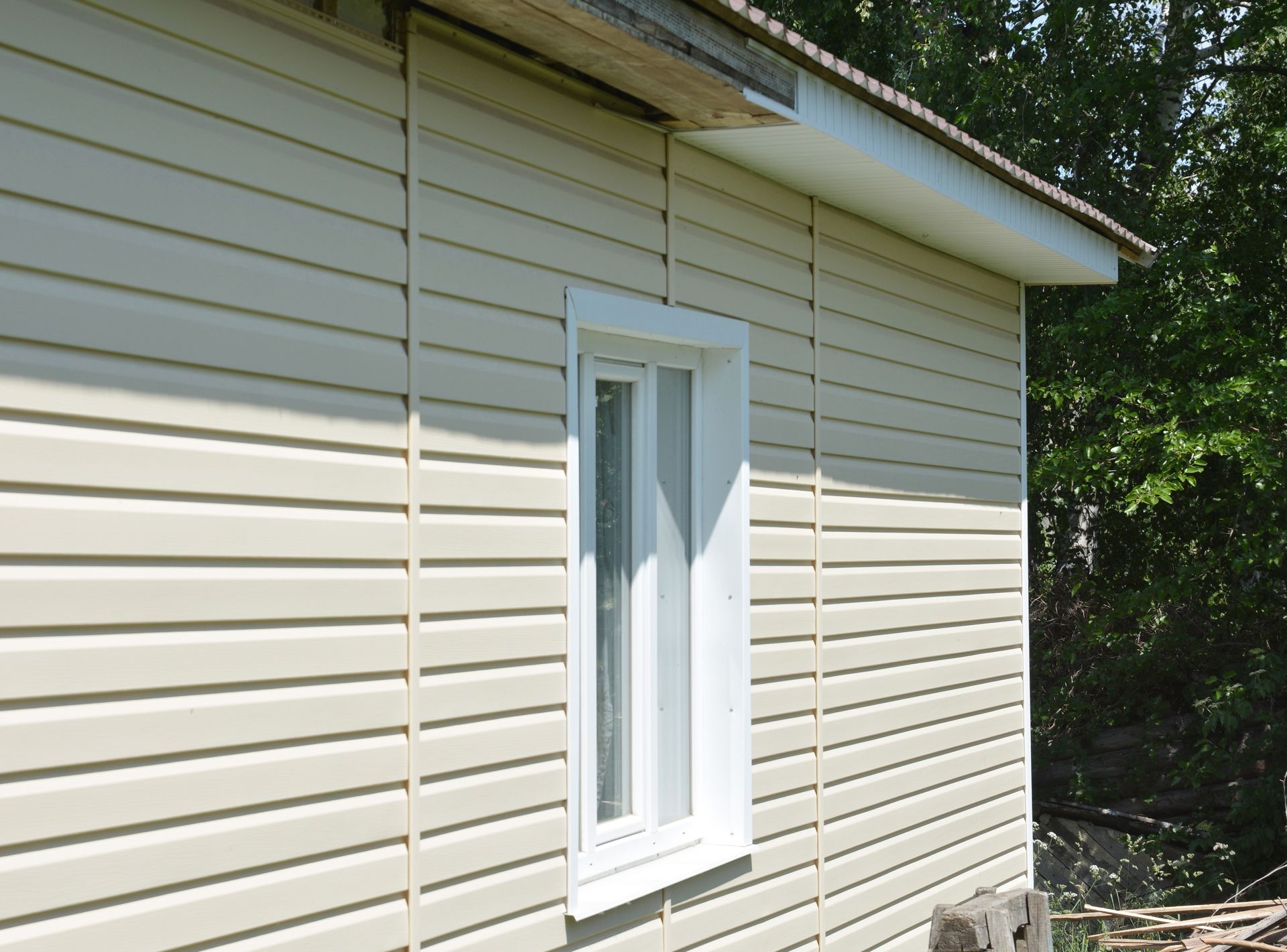A white-trimmed window set into a beige vinyl-sided exterior wall of a building near trees.