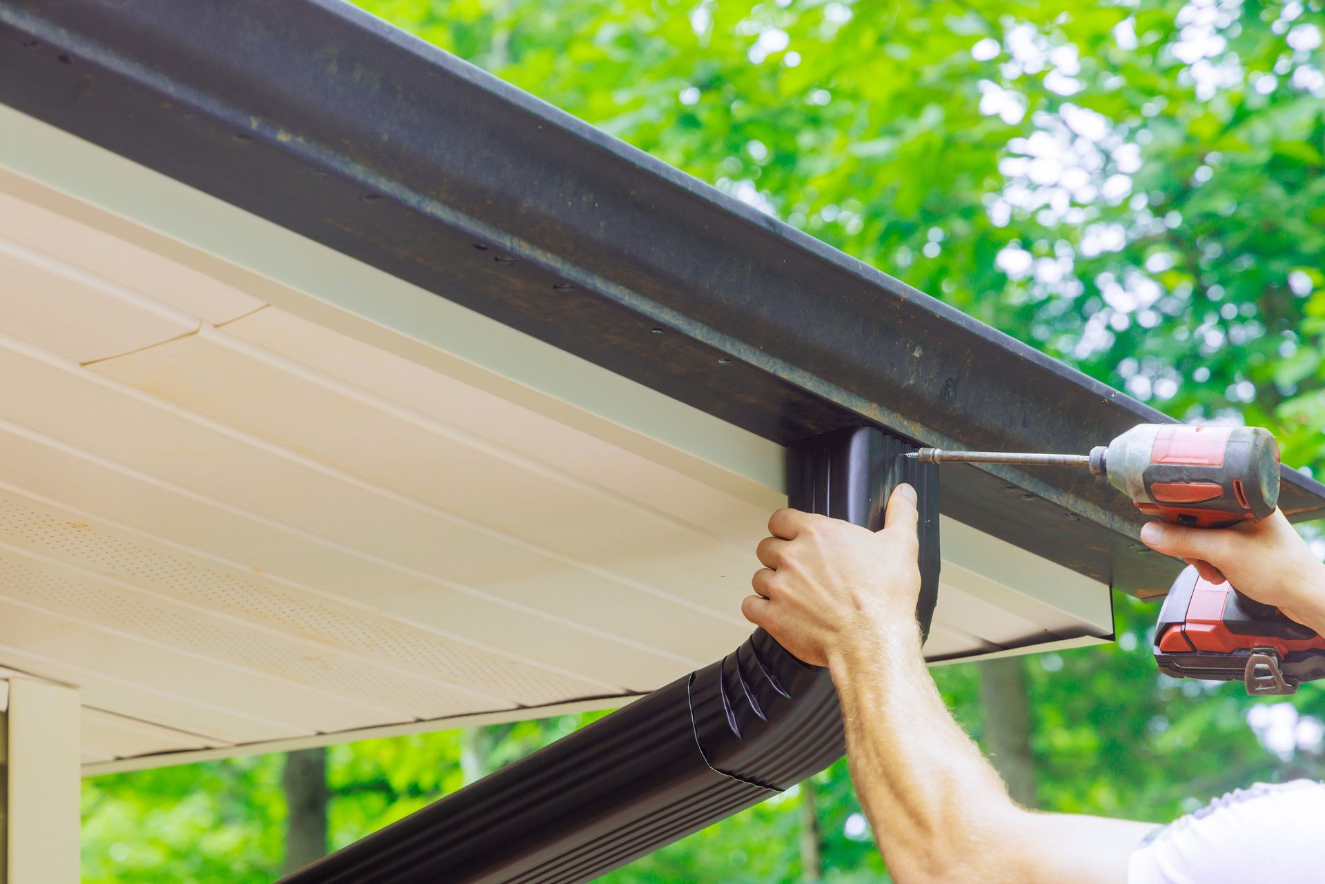 A person using a cordless drill to attach a dark brown gutter downspout to a roofline.