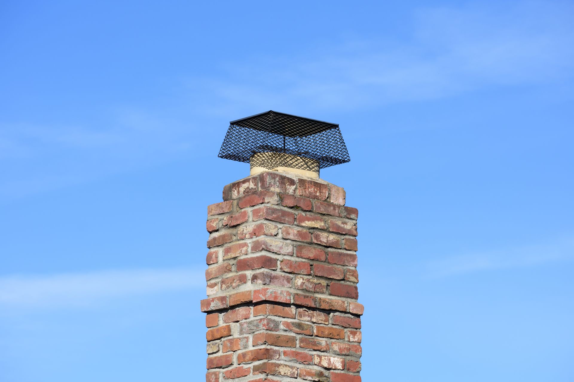 A brick chimney topped with a black wire mesh cap set against a clear blue sky.