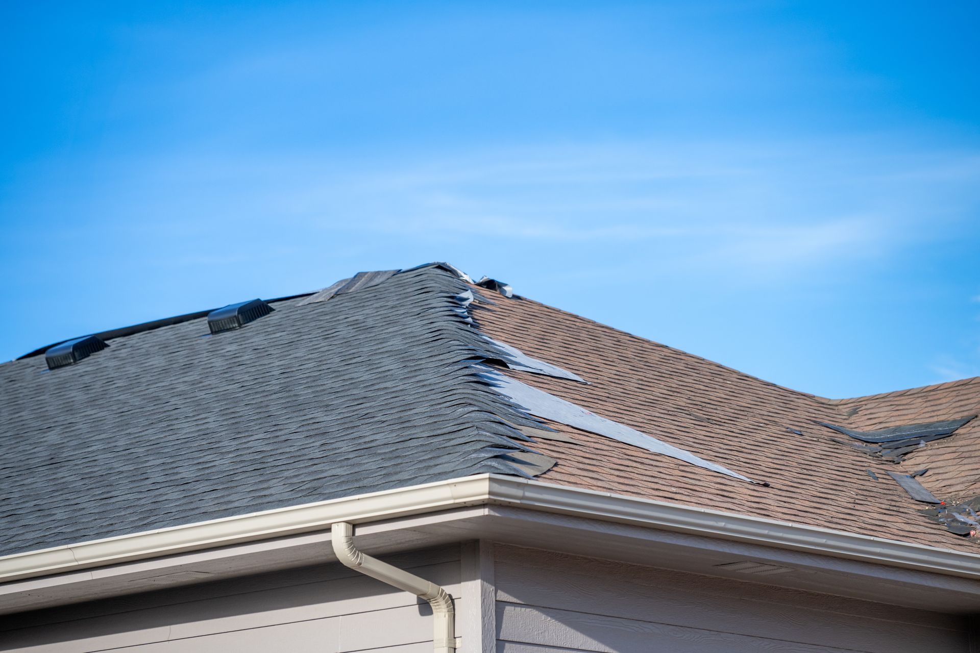 A residential roof with damaged, missing shingles along the hip ridge against a bright blue sky.
