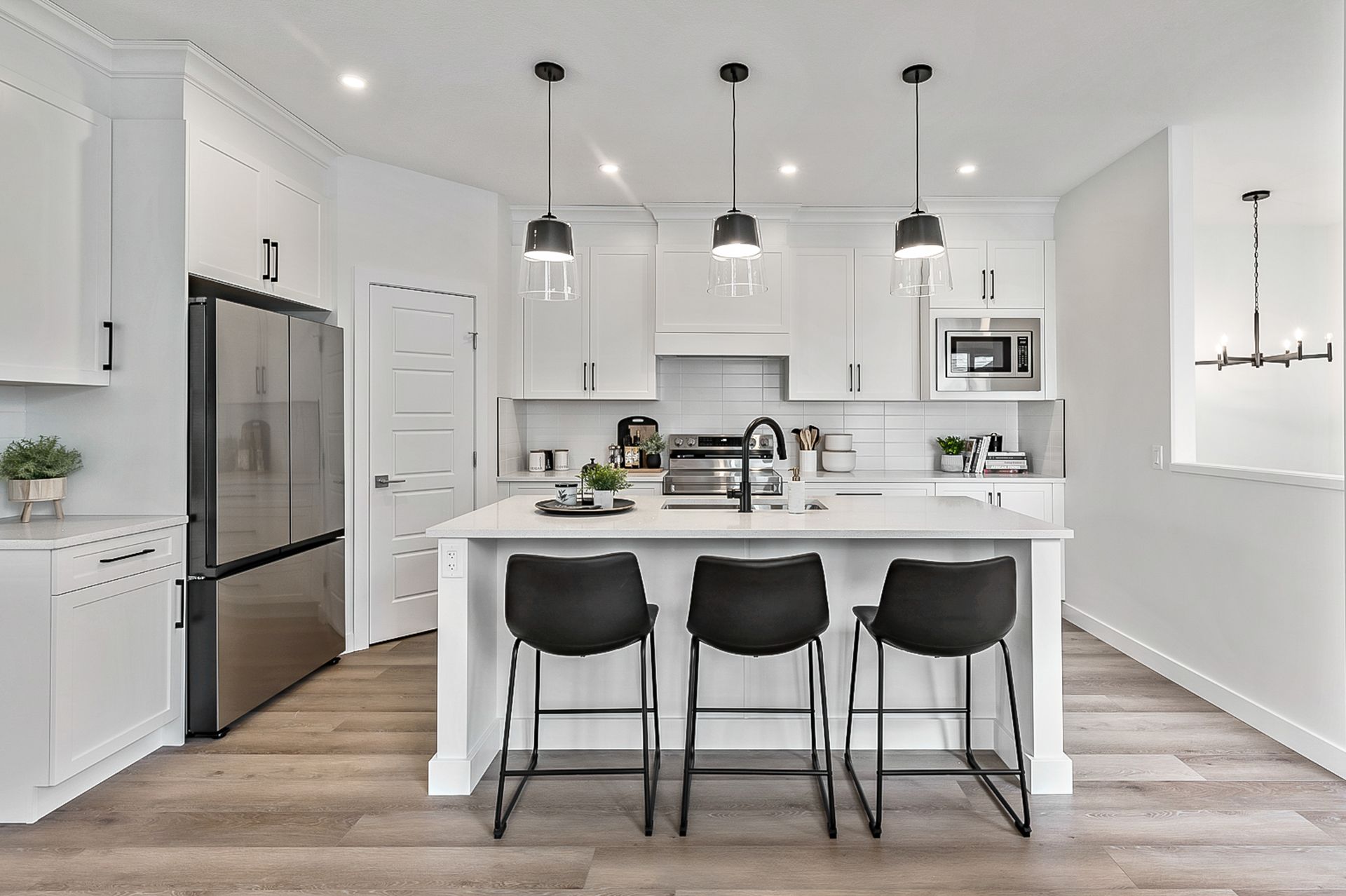 Modern white kitchen with island, black chairs, stainless steel appliances, wood floor.