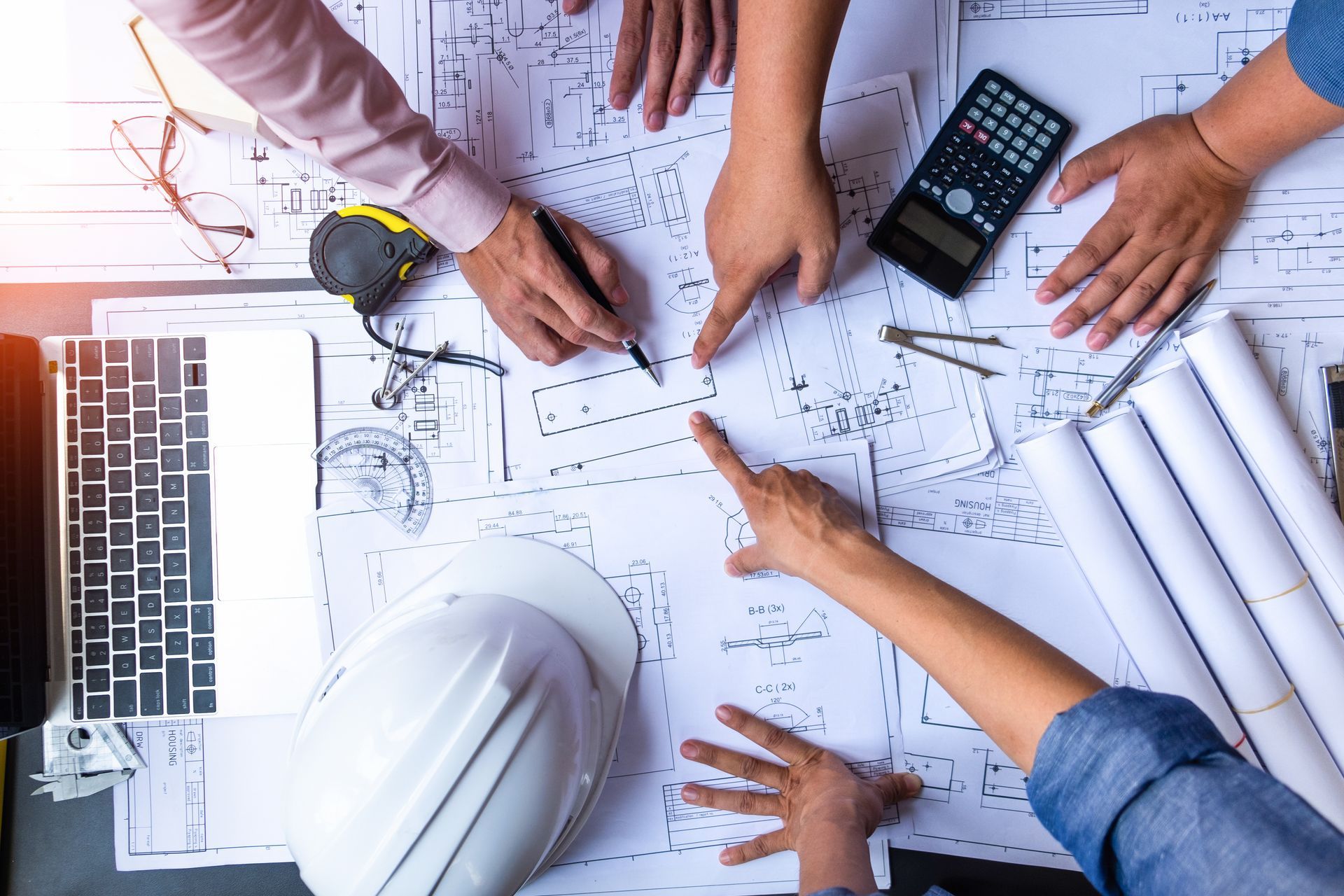 Hands pointing at blueprints on a table with a calculator, hard hat, and laptop.