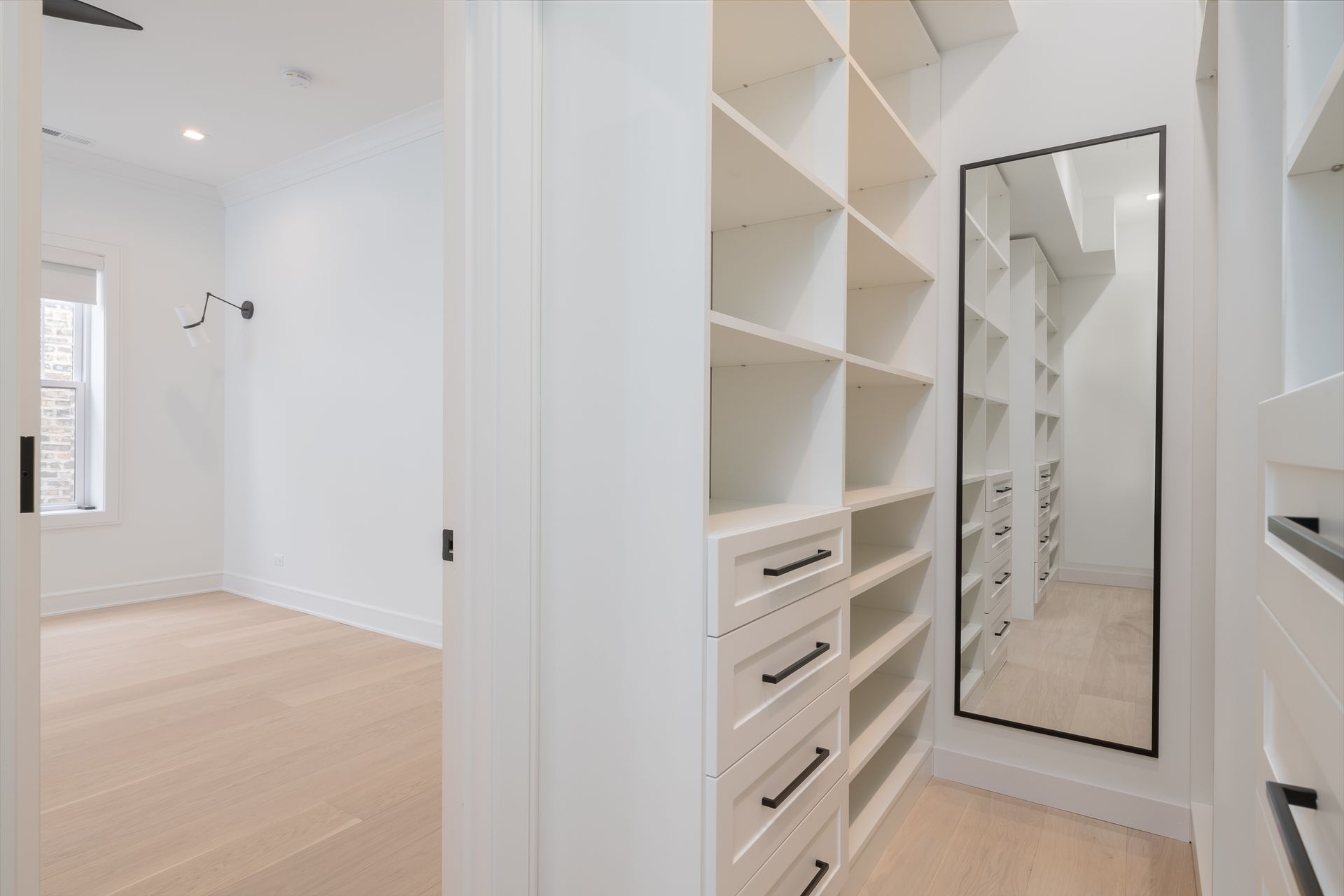 Walk-in closet with white shelving, drawers, and a large mirror. Light wood flooring.