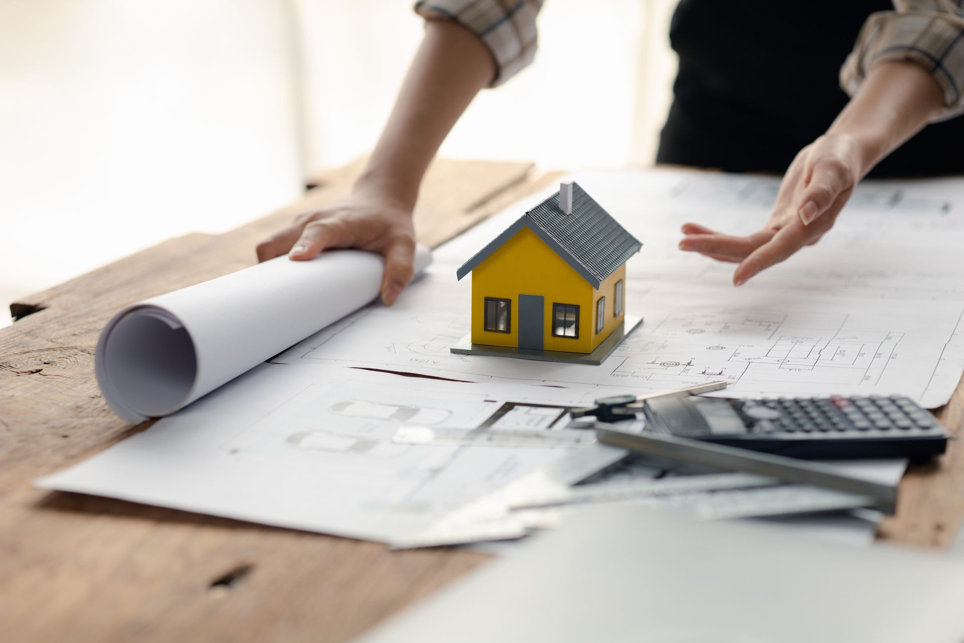 Person reviewing house plans with a model house, blueprints, and a calculator on a wooden table.