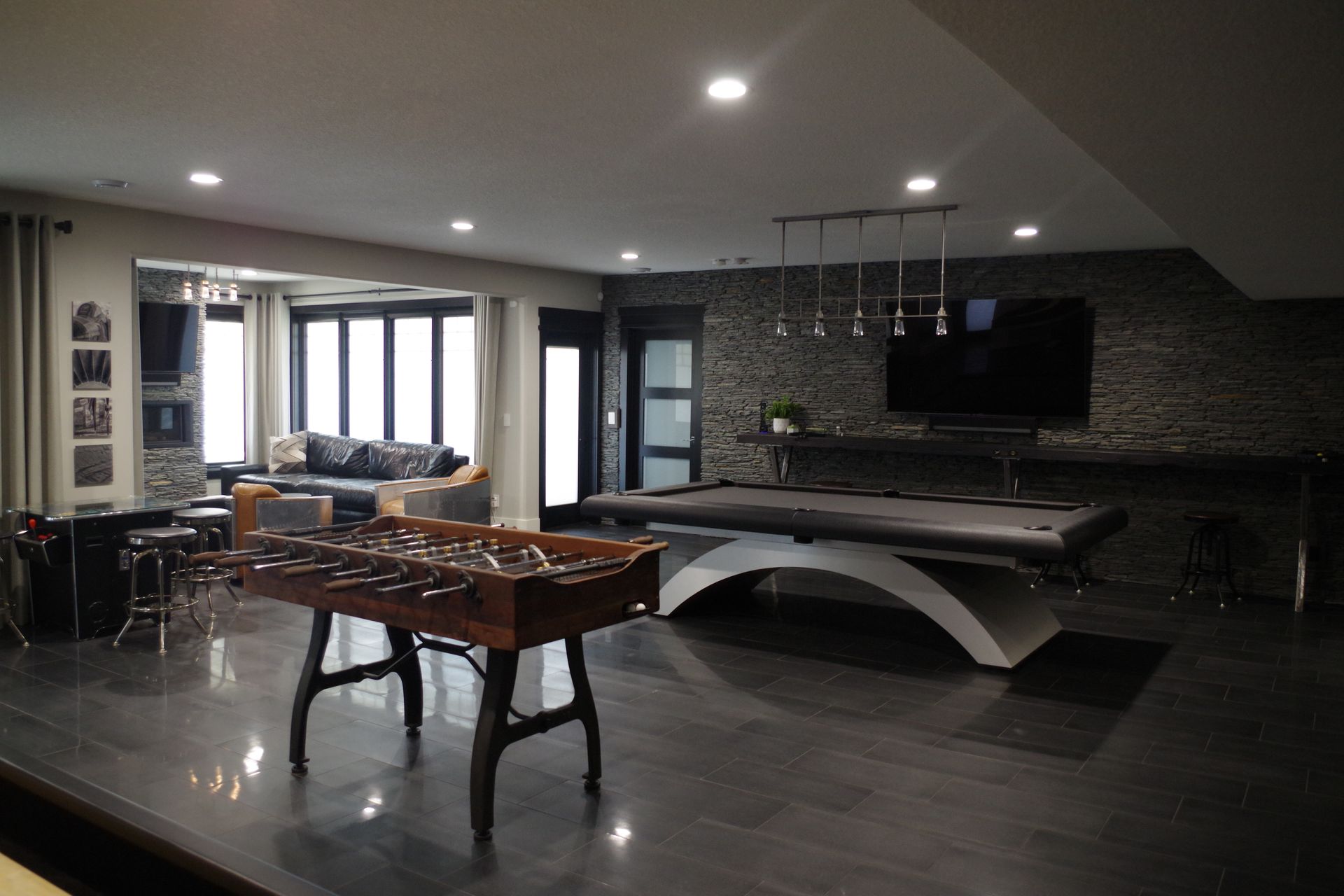 Game room with a foosball table, pool table, and TV against a dark stone wall.
