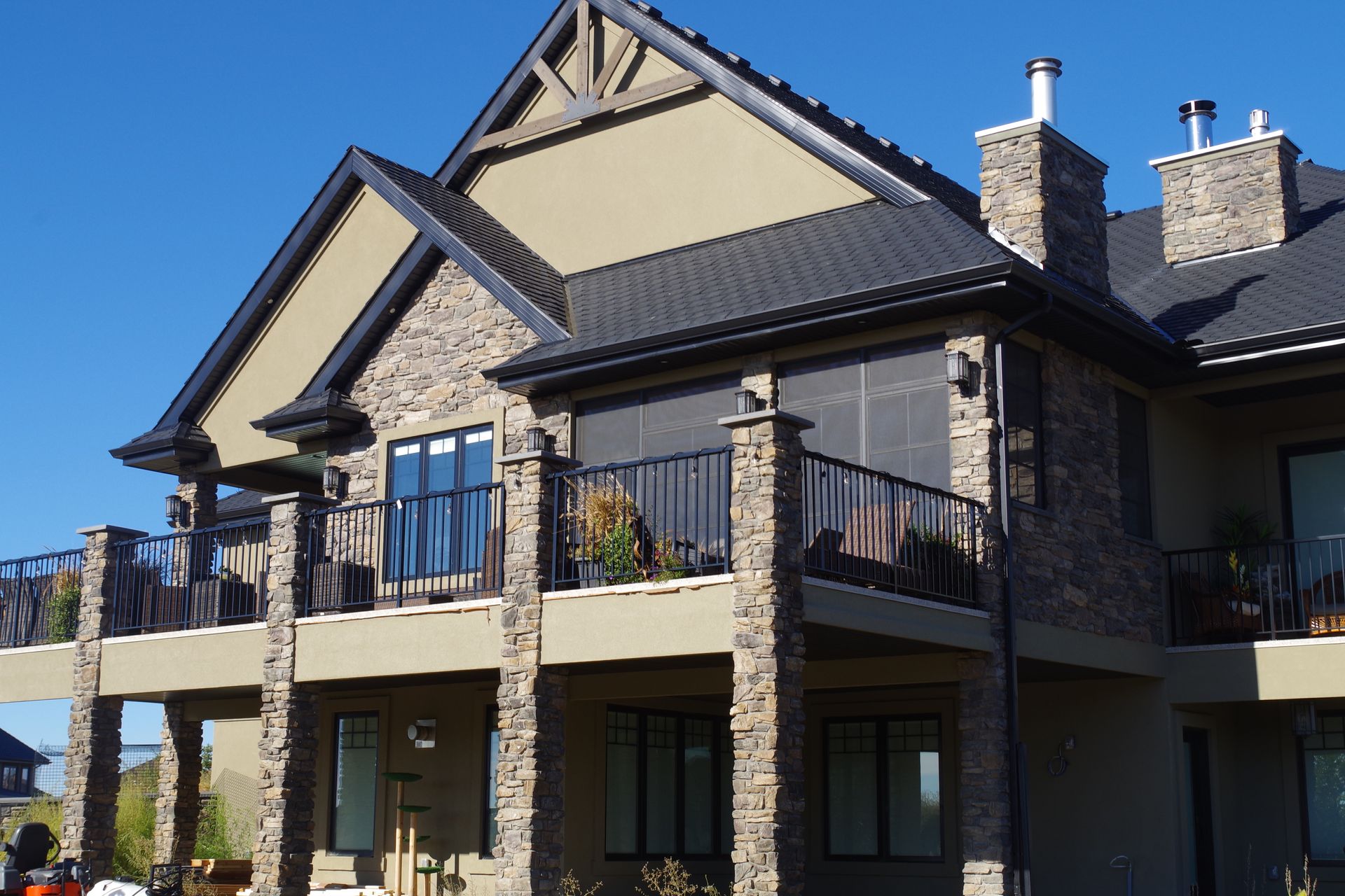 Multi-story stone and stucco home with balconies and dark roofing against a clear blue sky.