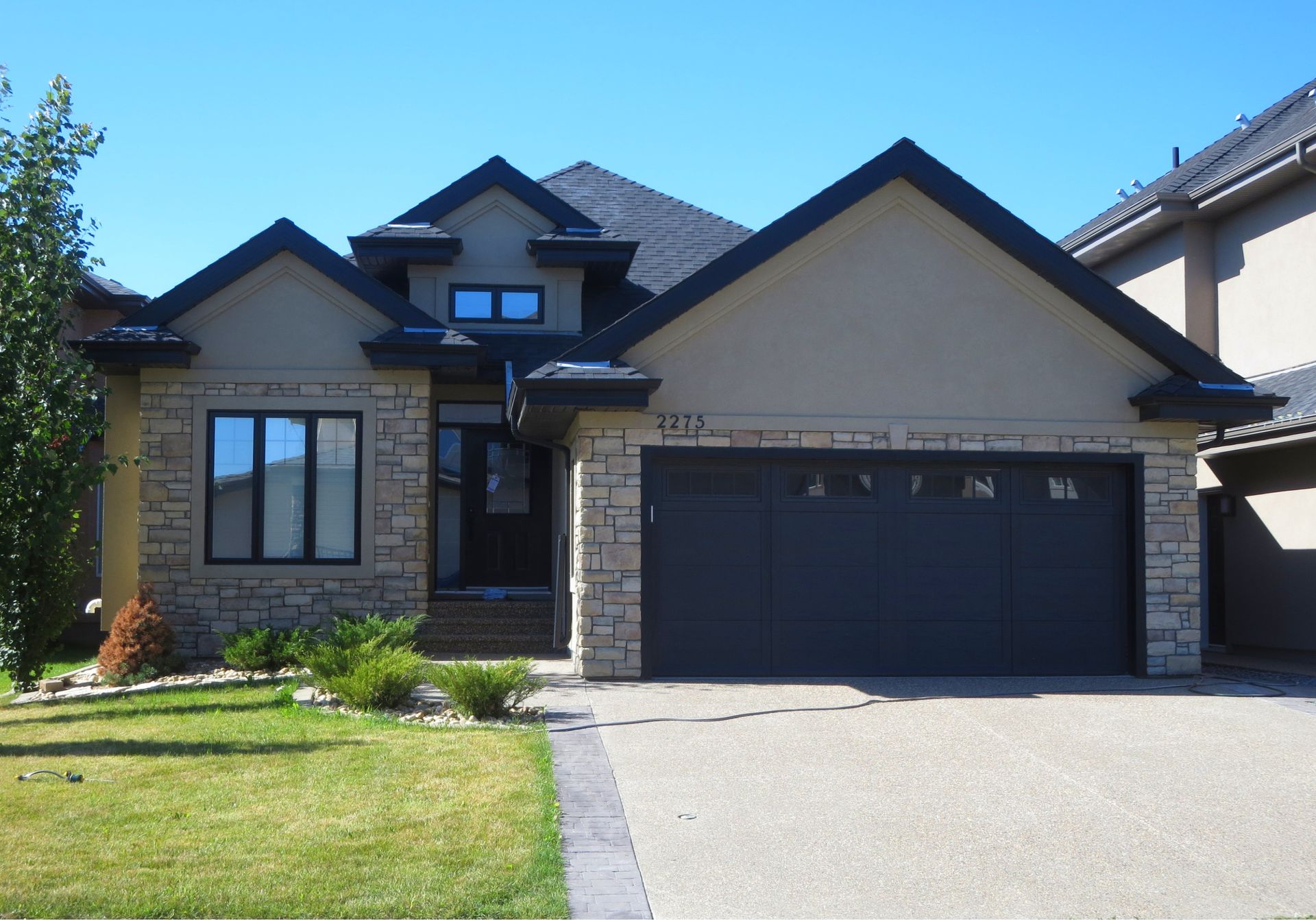 A two-story house with stone and tan siding, black garage door, and green lawn.
