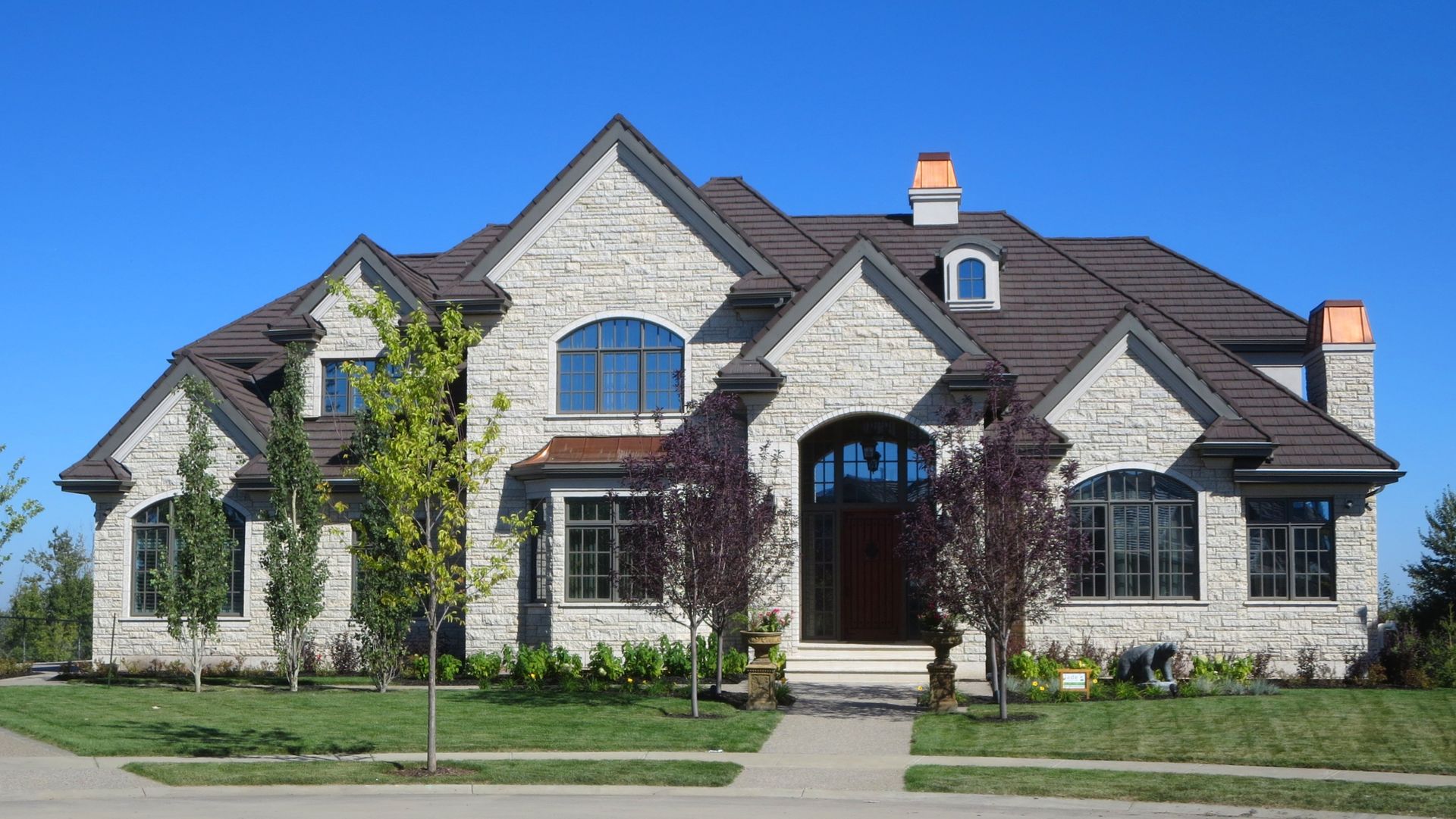 Two-story stone house with brown roof, arched windows, and front yard with trees against a blue sky.