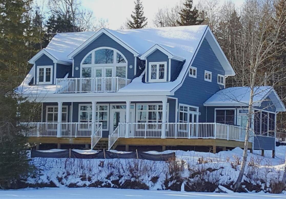 Blue house with a covered porch and snow-covered roof, near trees, with snowy ground in winter.