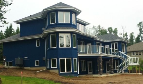 Blue house with multiple windows, deck, and glass railings, set amidst trees.