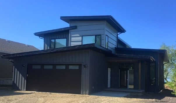 Modern two-story house with dark gray siding, black garage door, and large windows under a bright blue sky.